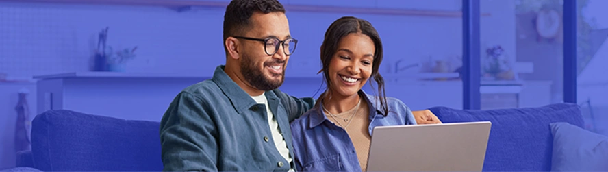 Um homem e uma mulher, sentados no sofá, usam o notebook e riem levemente.