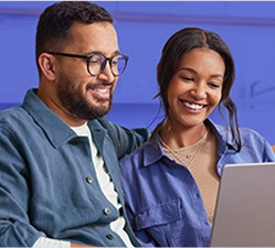 Um homem e uma mulher, sentados no sofá, usam o notebook e riem levemente.