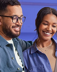 Um homem e uma mulher, sentados no sofá, usam o notebook e riem levemente.